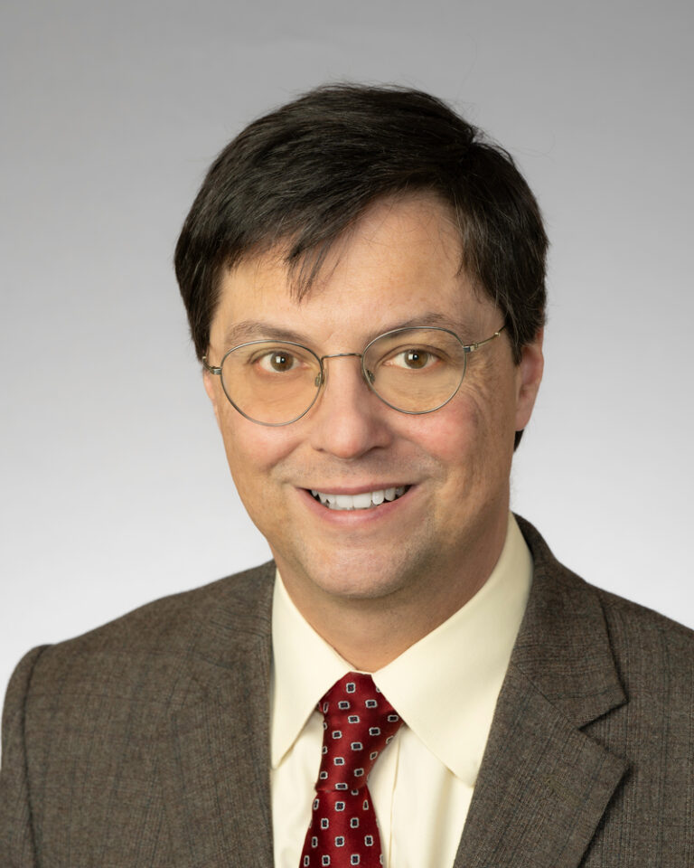 Dr. Leonard Appleman smiles directly at the camera in a professional headshot. He is in front of a white/grey plan photo backdrop, wearing wire-rimmed glasses, a brown-grey suit jacket, an offwhite, pale yellow button-down shirt, and a shiny deep red tie detailed with navy blue squares rimmed in gold that neatly decorates the red fabric in an alternating pattern. Dr. Appleman has white skin and short dark brown hair combed in a plain, professional style. His warm brown eyes crinkle slightly as he smiles.