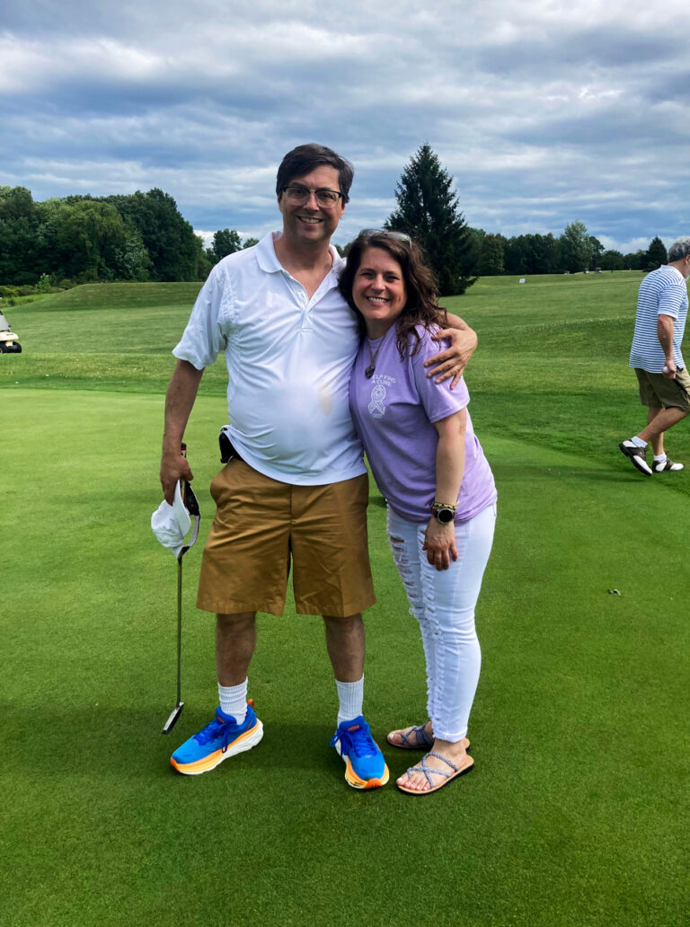 Dr. Leonard Appleman wraps his left arm around, Gail Tucci, as they stand side-by-side on a golf course. There is part of a golf cart in the distance on the left and a man in a powder blue polo walking away on the right. It is a partly cloudy day as everyone stands on vibrant green golf turf lined with full-leafed dark green deciduous and coniferous trees in the far background. Dr. Appleman wears 1950s black browline-styled glasses, a white golf polo, khaki shorts, and bright royal blue and orange tennis shoes with white high socks. He holds a golf club in his right hand, aimed toward the green. Gail is on Dr. Appleman’s left side, with shoulder-length brown hair that is gently curled. She wears a lavender t-shirt with a white decal on the upper left quadrant of the shirt, light blue skinny jeans with distressed details on the front, and strappy slip-on sandals. Both Dr. Appleman and Gail are smiling widely at the camera.