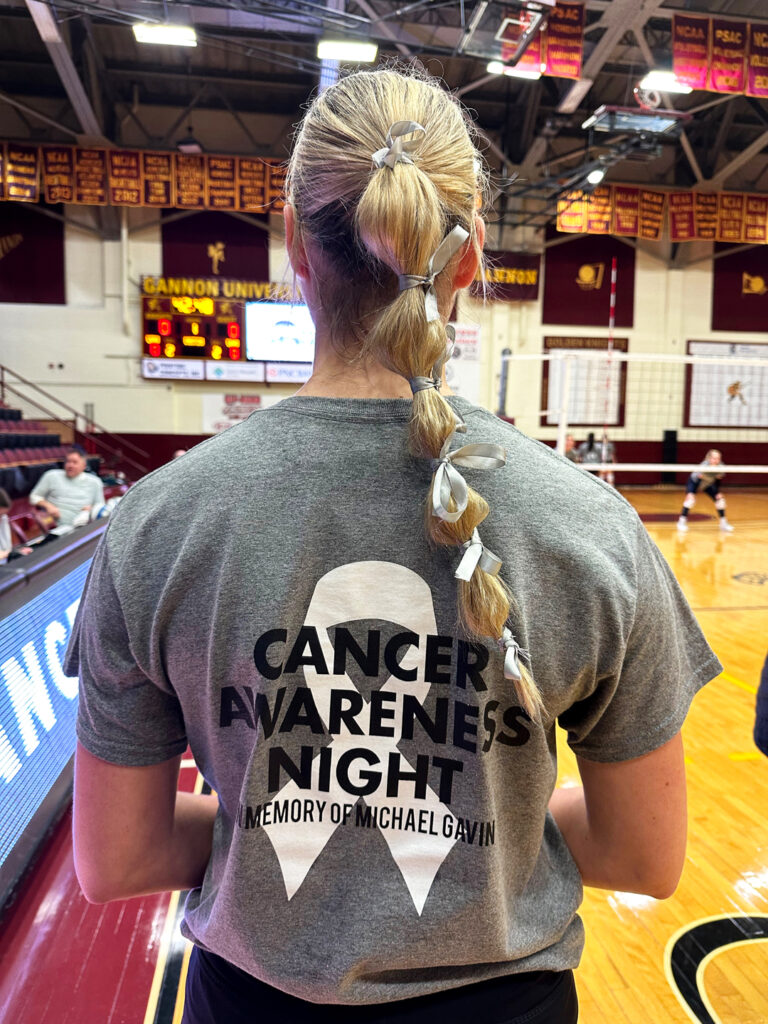 Mekenna Gavin, wearing a grey t-shirt with a white ribbon decal and the words “Cancer Awareness Night, Remembering Michael Gavin,” and her long blonde hair tied in a ponytail with white ribbons, looks away from the camera to observe the Gannon gymnasium court where she organized a volleyball game to honor her late father, Michael Gavin.