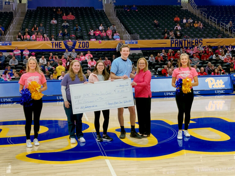 Four people - 3 women and 1 man - stand together smiling in the middle of the University of Pittsburgh’s basketball court during halftime. They hold a large white check (about 2x4 feet) with a donation amount on it. Two young girls from the Pitt dance team stand on either side of the group, wearing pink shirts and black leggings, smiling and holding blue and gold pompoms. There is a crowd of spectators for the basketball game sitting in the stands in the background. A yellow banner hangs above them with “The Zoo” and “PANTHERS” written in dark blue letters.