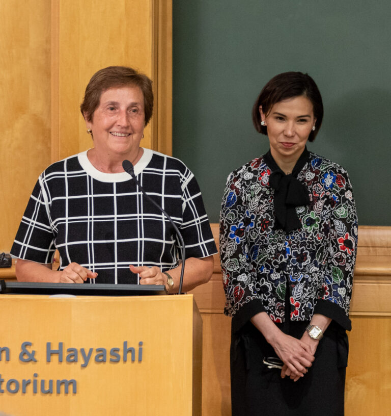 Two women standing at a podium, one wearing a black and white checkered shirt, smiling as she speaks; beside them stands another woman dressed in a stylish business casual outfit with a dark brown bob.