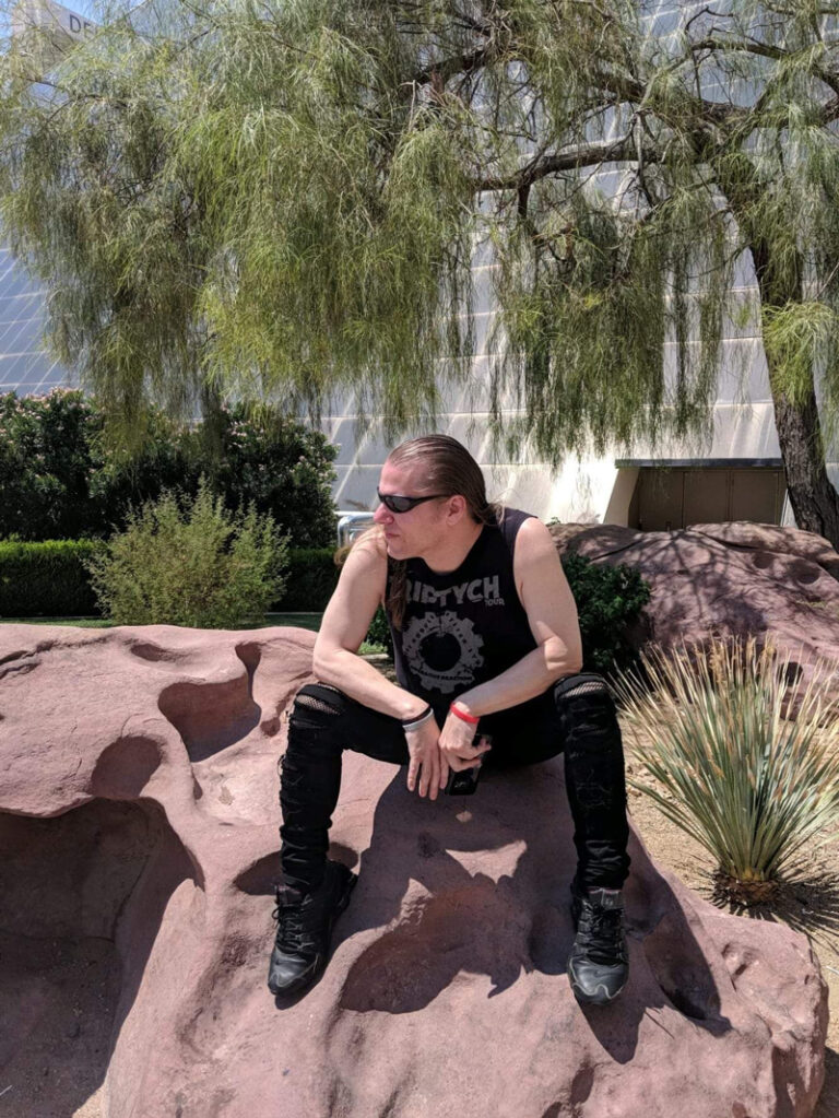 Jim Semonik sits stoically on a reddish tan rock, looking off in the distance with his head turned to his right and his arms resting casually on his knees. He is surrounded by spiky green succulents next to him and a willow-like green tree in the background. Jim is donning all black in a cool, alternative style. He wears a black graphic tank top that is loose with distressed white graphics, black distressed skinny jeans, and all-black high-top athletic shoes. He completes his look with black sunglasses and white, black, and red bracelets.