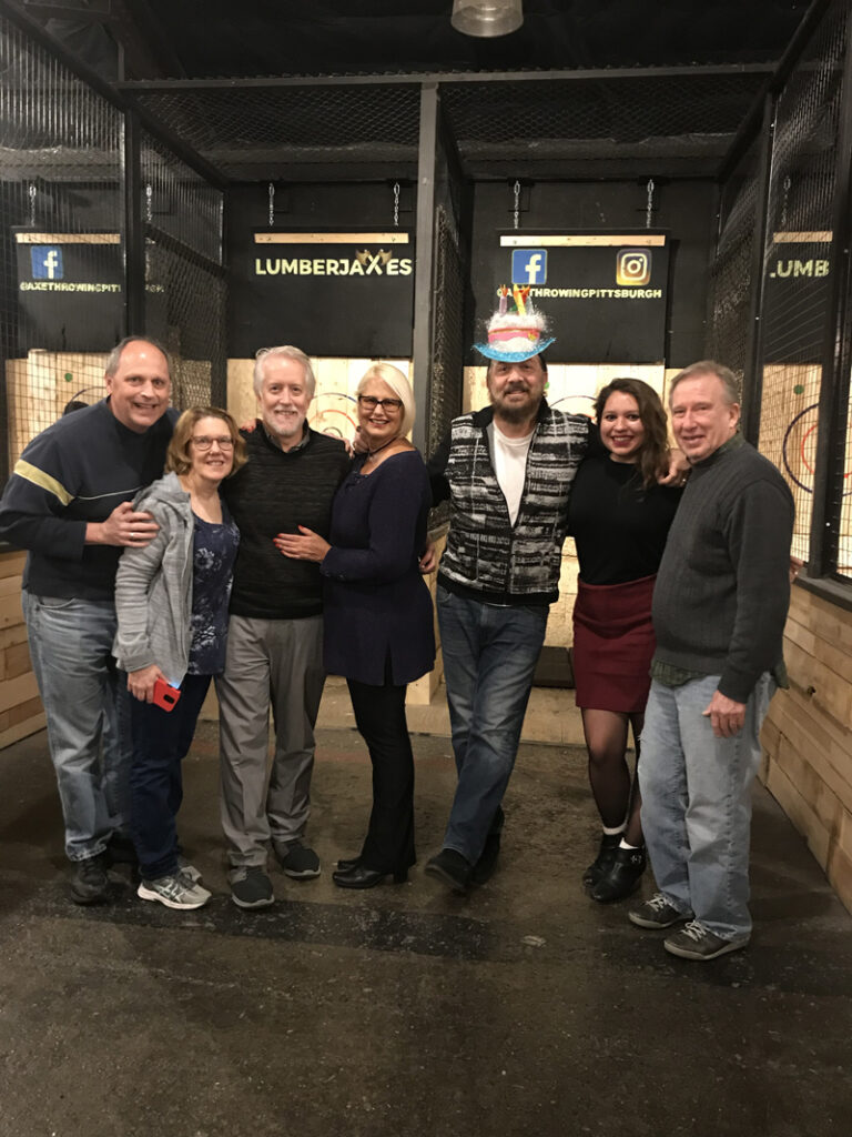 A group of 7 white adults stand together in a row as they interlock arms and smile warmly for the camera. They are standing in front of a wall of wooden targets in an industrial-styled warehouse - a local business called LumberJaxes in Pittsburgh, PA. LumberJaxes’ logo is displayed on the far wall above the adults’ heads, in a yellow plain font on a black wall. The group looks to be celebrating a birthday as one adult in the middle of the group wears a large, wide-brimmed plush hat in the shape of a white birthday cake with multicolored candles.