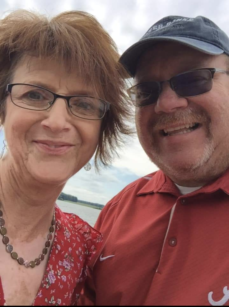 Close-up of Kay Boyd Rowe and her husband, Robin. Both are grinning as they stand side by side for a selfie-styled photo on a sunny day. In the background, it looks like they are at an ocean or lake as there is a glimpse of water in the background. Kay, looking to be in her 50s-60s, is on the left of the frame with short, spikey brown hair and bangs. She is wearing rectangular wire-framed black prescription glasses, a red floral v-neck shirt, and a copper-colored necklace comprised of circular flat beads around her neck. Robin, on the right of the frame, also looks to be in his 50s-60s with white-gray stubble on his face and neck. He is wearing a worn dark blue ballcap, rectangular transitional prescription glasses, and a red Nike polo shirt.