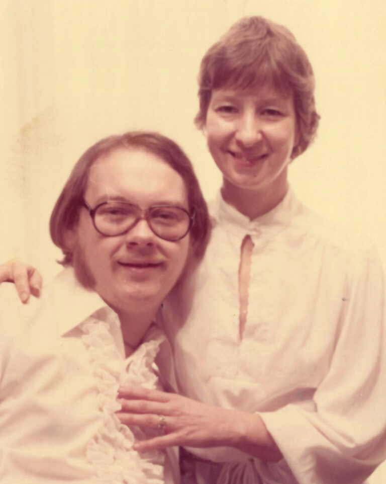 Two people, a woman and a man smiling for the camera in a vintage photograph. Both are in white shirts and have short light brown hair. The man is sitting and wearing glasses as the woman is standing on his left with her arms around him.