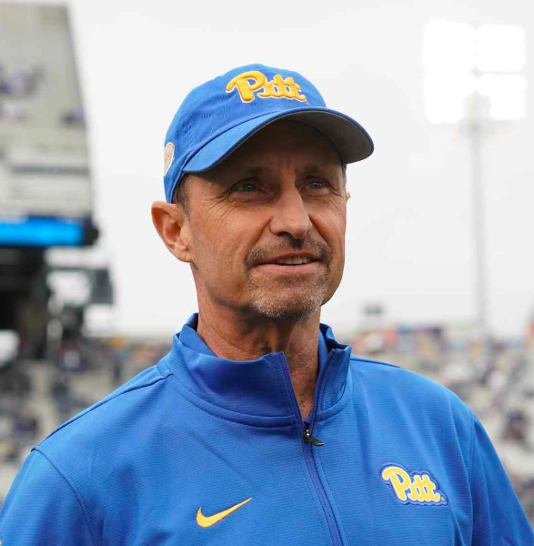 Pittsburgh Panthers Coach Randy Bates wearing a University of Pittsburgh ball cap and zip-up while smiling cheerfully and watching a football game off-frame.